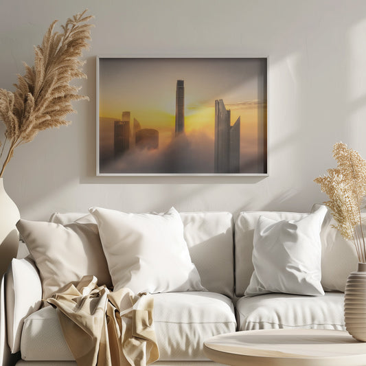A framed photograph of a modern city skyline with skyscrapers emerging from a thick layer of clouds at sunrise, creating a dramatic and ethereal scene against a golden sky. Wall Art