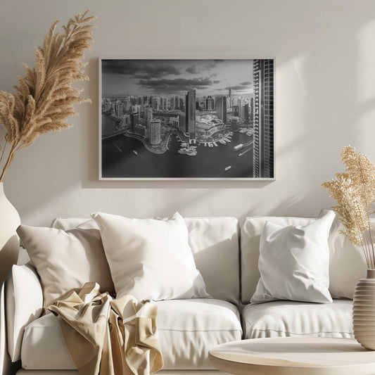 A framed, black and white panoramic photograph of the Dubai Marina taken from a high vantage point. The image captures the dense cityscape with numerous modern skyscrapers surrounding a winding waterway filled with docked yachts and moving boats. The sky is filled with dramatic, dark clouds, adding to the moody atmosphere of the urban landscape. Decor