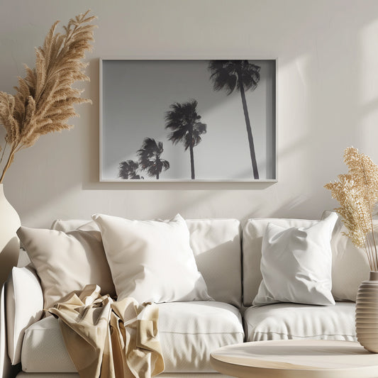A framed black and white photograph of four silhouetted palm trees of increasing height, arranged diagonally against a plain, light grey sky. The palm fronds are blurred, suggesting they are blowing in the wind. Decor