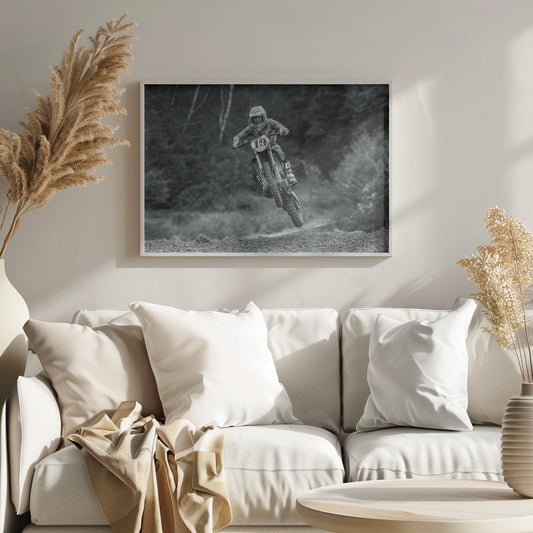 A dynamic black and white photograph of a motocross racer in mid-air, jumping over a dirt mound. The rider, wearing full protective gear and a helmet, is on a dirt bike with the number 19 plate. The background is a soft-focus forest, and the entire image is enclosed in a silver frame. Poster