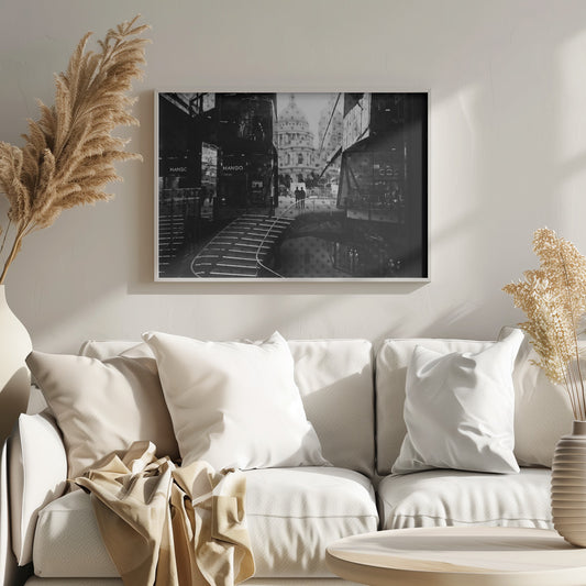 A framed, black and white photograph capturing a layered urban scene. The view, seen through a window with reflections of a curved, lit staircase, shows two people walking towards a large domed cathedral framed by modern glass buildings. Wall Art
