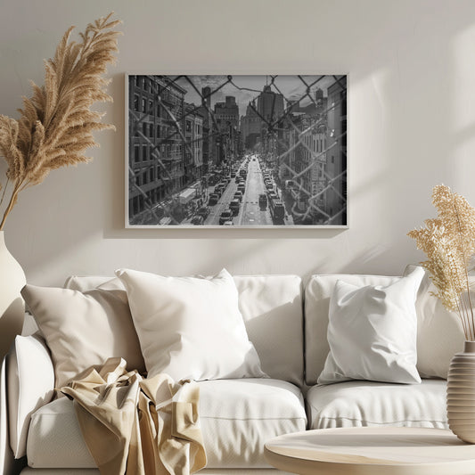 A high-angle, black and white photograph of a bustling New York City street viewed through a chain-link fence. The fence is in the foreground, framing the scene of traffic-filled streets, historic buildings with fire escapes, and skyscrapers in the distance under a cloudy sky. Wall Art