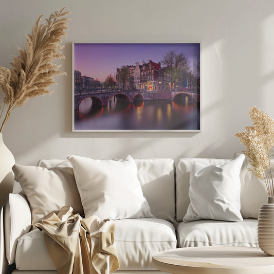 A scenic view of an Amsterdam canal at twilight, with historic brick buildings and illuminated arched bridges reflected in the calm water under a vibrant pink and purple sky. Poster
