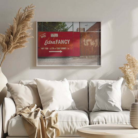 A framed photograph of a bright red outdoor sign for a restaurant named 'Extra Fancy'. The sign, painted with white retro-style text, advertises hot food and cold drinks and is situated next to a rusty, corrugated metal fence on a city sidewalk. Artwork