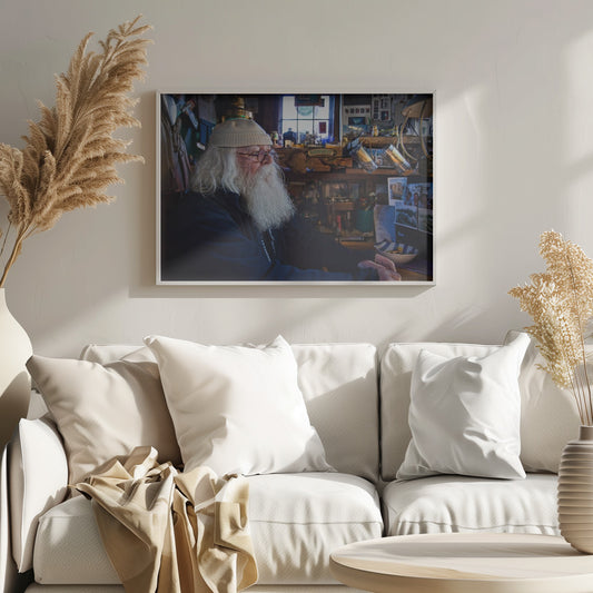 A profile portrait of an elderly man with a long, full white beard and hair, wearing glasses and a cream-colored knit beanie. He sits indoors in a warmly lit, cluttered room that looks like a cabin or a pub, with his hands clasped in front of him, gazing thoughtfully to the side. Poster