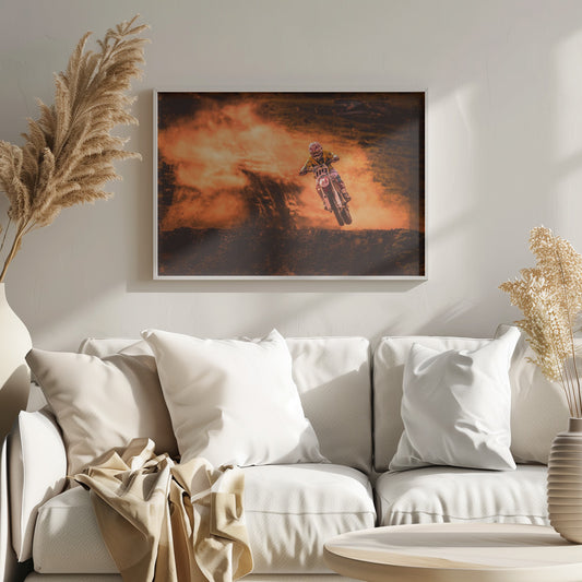 An action-packed photo of a motocross rider in mid-air, jumping their dirt bike over a mound of earth. A large cloud of orange dust billows behind them, creating a dramatic and fiery background. Wall Art