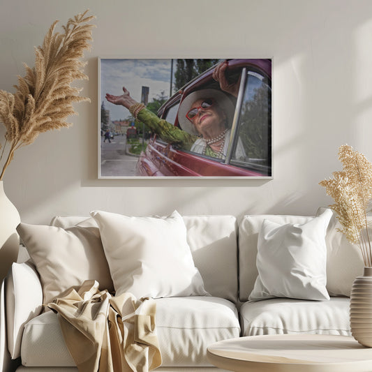 A stylish elderly woman wearing a white hat, red sunglasses, and pearl necklaces leans out of the passenger window of a classic red car. Her arm is outstretched with a joyful expression on her face, enjoying a ride through a city street. Poster