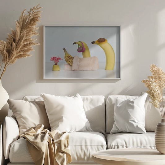 A whimsical still life photo featuring three bananas personified as a family or friends. One overripe, spotted banana lies down, covered by a pink towel, while another banana with a tiny pink purse and a third with a small hat seem to be visiting. A small vase with pink flowers sits nearby on the white surface. Poster