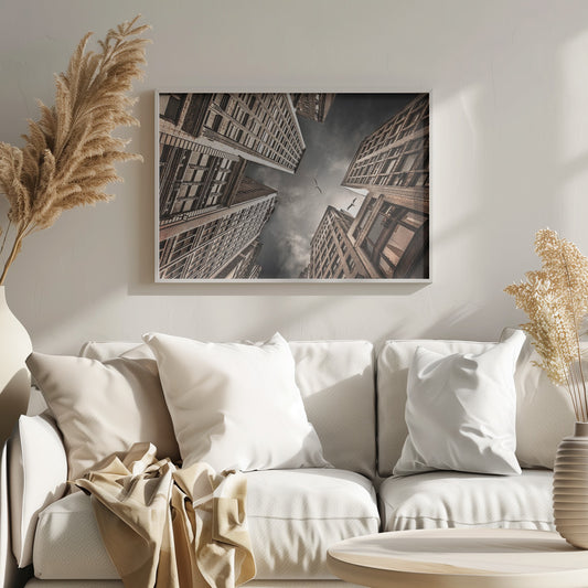 A dramatic, low-angle photograph looking up at the sky between several tall, sepia-toned skyscrapers that converge towards the center. Two birds are seen flying in the moody, dark grey sky. The image has a vintage feel and is presented in a silver frame. Wall Art
