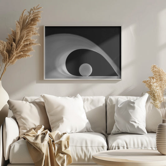 A dramatic black and white still life photo featuring a golf ball set against a stark black background. A large, abstract white shape swirls gracefully over the ball, creating a dynamic, wave-like composition. The image is presented within a silver frame. Decor