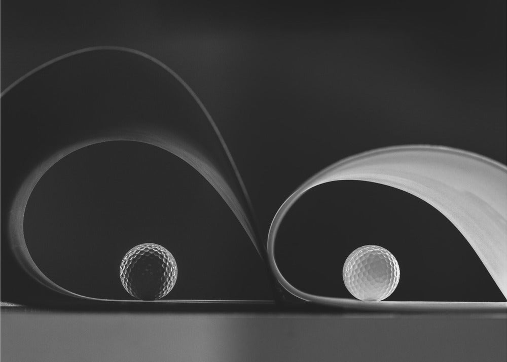 A framed, black and white abstract photograph featuring two golf balls nestled within the graceful curves of a single sheet of paper, creating a striking interplay of light, shadow, and form against a dark background. Decor