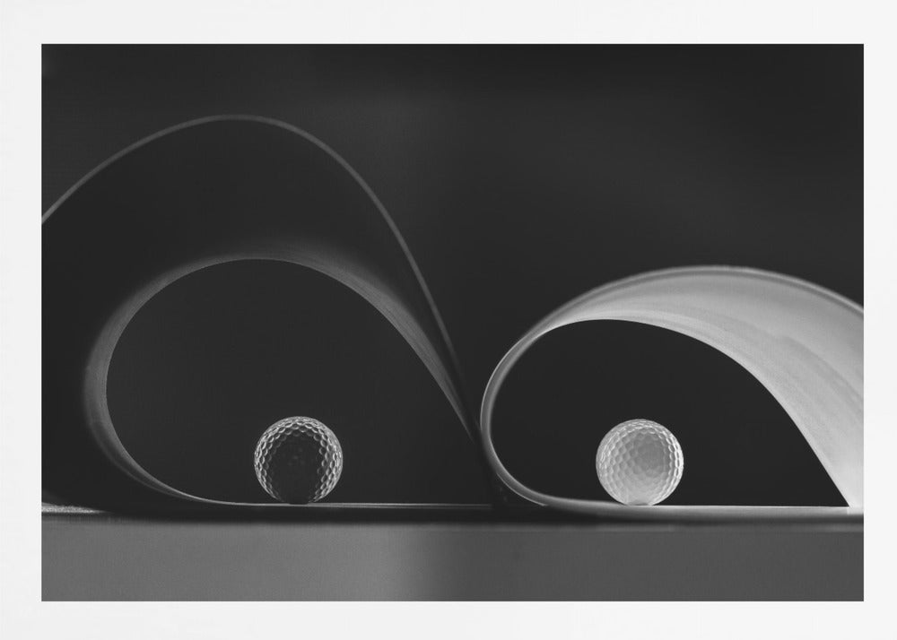 A framed, black and white abstract photograph featuring two golf balls nestled within the graceful curves of a single sheet of paper, creating a striking interplay of light, shadow, and form against a dark background. Decor