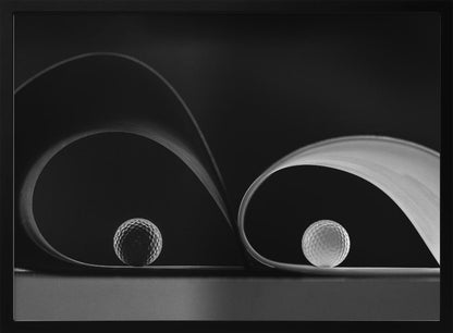 A framed, black and white abstract photograph featuring two golf balls nestled within the graceful curves of a single sheet of paper, creating a striking interplay of light, shadow, and form against a dark background. Decor