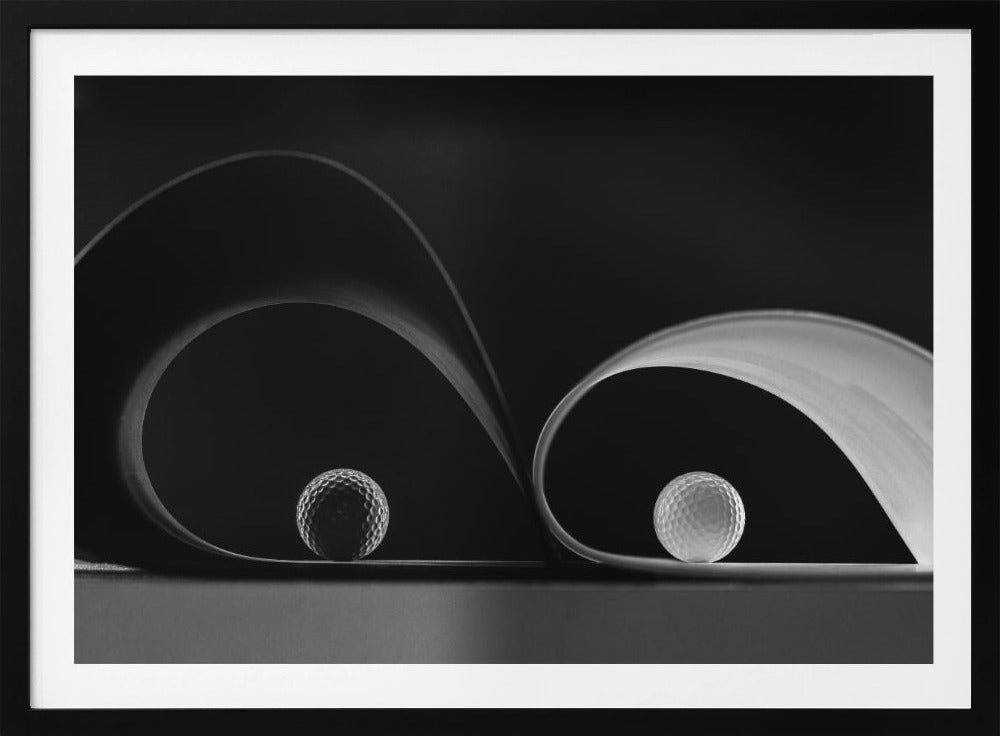 A framed, black and white abstract photograph featuring two golf balls nestled within the graceful curves of a single sheet of paper, creating a striking interplay of light, shadow, and form against a dark background. Decor