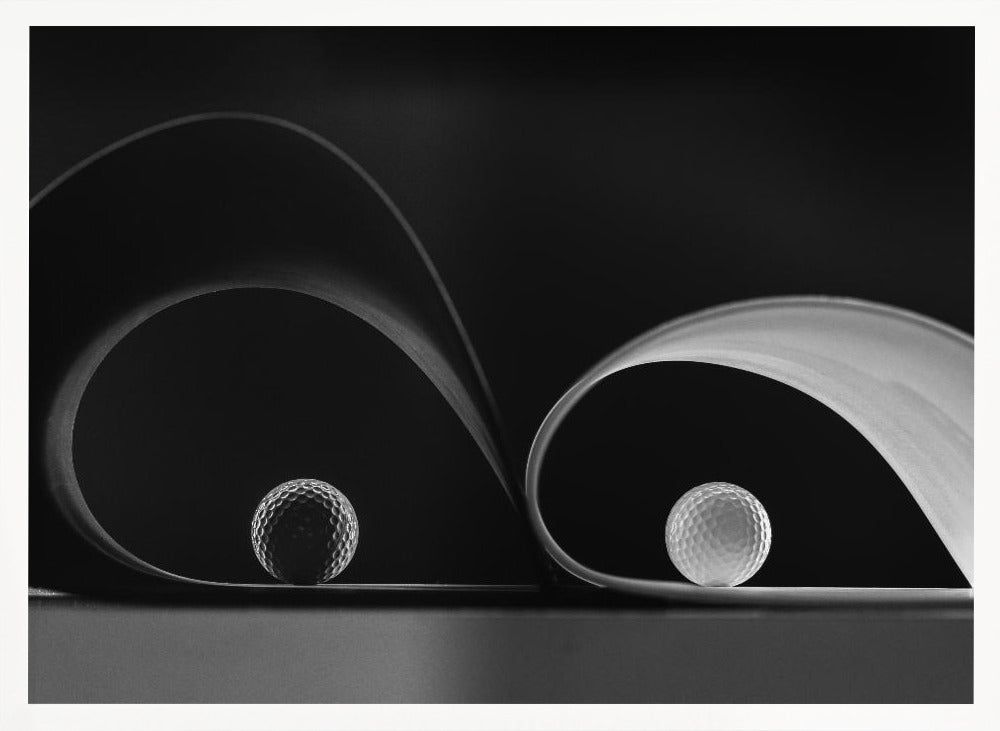 A framed, black and white abstract photograph featuring two golf balls nestled within the graceful curves of a single sheet of paper, creating a striking interplay of light, shadow, and form against a dark background. Decor