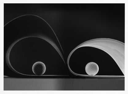 A framed, black and white abstract photograph featuring two golf balls nestled within the graceful curves of a single sheet of paper, creating a striking interplay of light, shadow, and form against a dark background. Decor