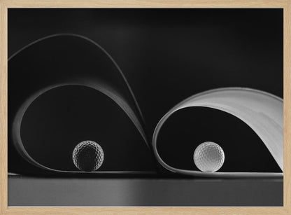A framed, black and white abstract photograph featuring two golf balls nestled within the graceful curves of a single sheet of paper, creating a striking interplay of light, shadow, and form against a dark background. Decor