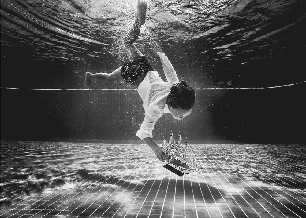A surreal black and white underwater photograph of a child in a white shirt and shorts diving towards the bottom of a tiled pool to retrieve a miniature model of a sailing ship. Artwork