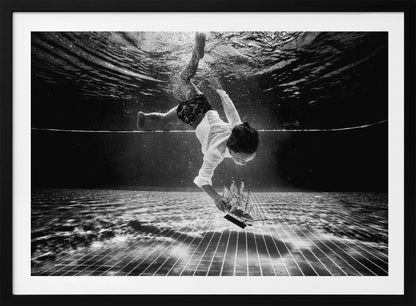 A surreal black and white underwater photograph of a child in a white shirt and shorts diving towards the bottom of a tiled pool to retrieve a miniature model of a sailing ship. Artwork