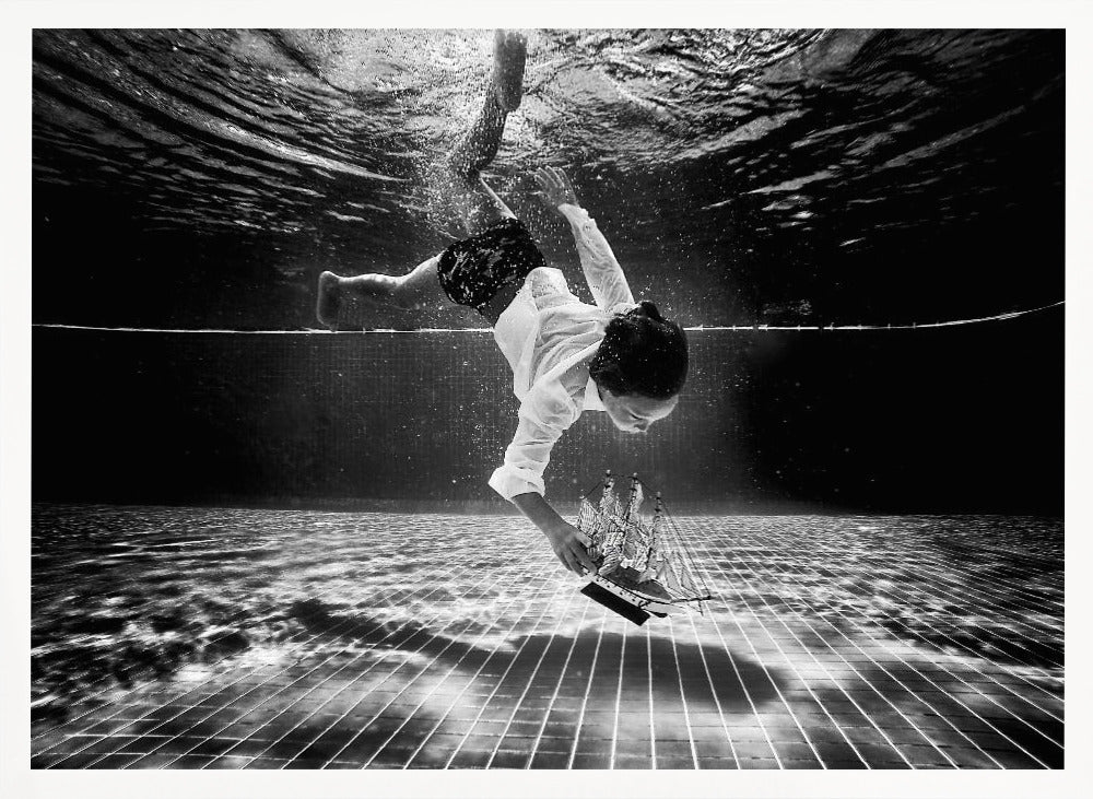 A surreal black and white underwater photograph of a child in a white shirt and shorts diving towards the bottom of a tiled pool to retrieve a miniature model of a sailing ship. Artwork