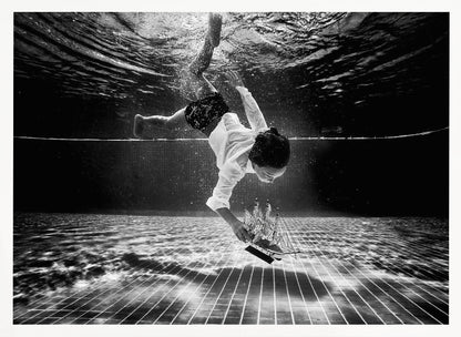 A surreal black and white underwater photograph of a child in a white shirt and shorts diving towards the bottom of a tiled pool to retrieve a miniature model of a sailing ship. Artwork