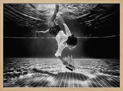 A surreal black and white underwater photograph of a child in a white shirt and shorts diving towards the bottom of a tiled pool to retrieve a miniature model of a sailing ship. Artwork