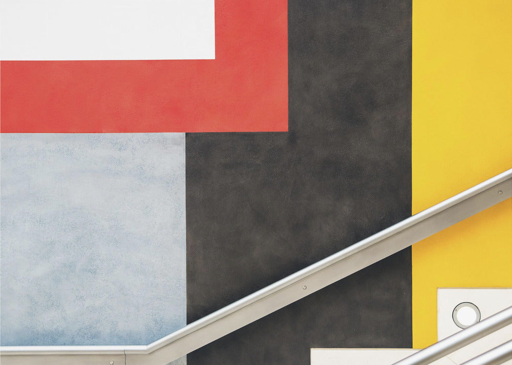 A framed abstract art piece featuring a composition of large, solid-colored geometric shapes in red, yellow, black, white, and light gray. A silver handrail cuts diagonally across the lower portion of the image, adding a sharp line and a sense of architectural space. Artwork