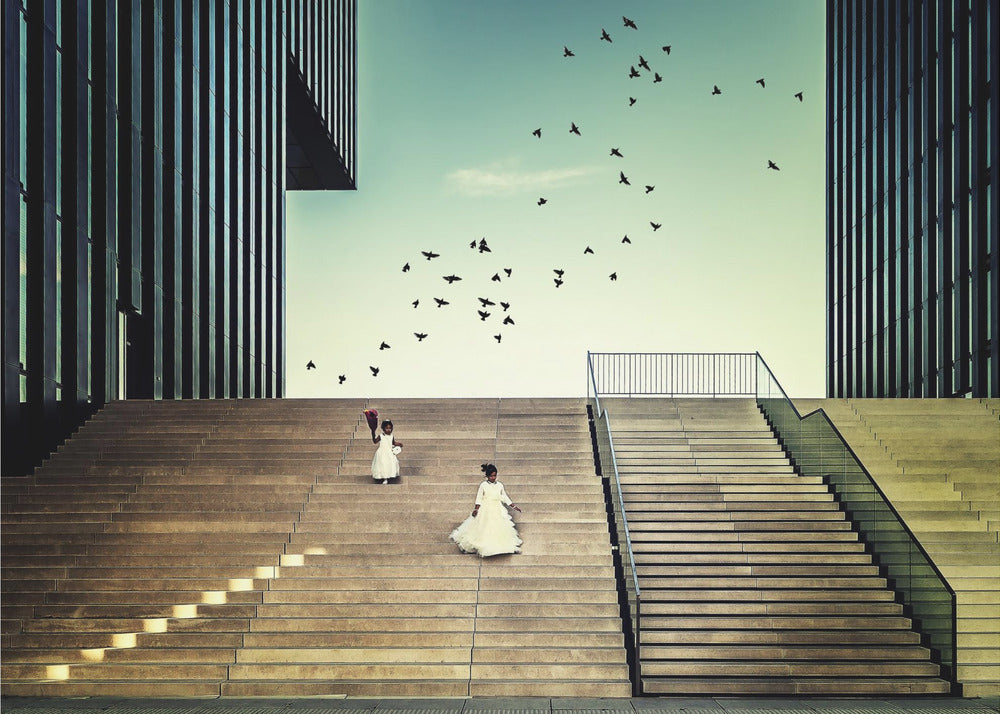 A framed piece of art depicting two young girls in white dresses climbing a vast, modern concrete staircase flanked by two towering, dark buildings. A large flock of birds flies through the teal sky above them, adding a sense of movement to the structured, architectural scene. Poster
