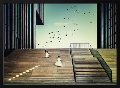A framed piece of art depicting two young girls in white dresses climbing a vast, modern concrete staircase flanked by two towering, dark buildings. A large flock of birds flies through the teal sky above them, adding a sense of movement to the structured, architectural scene. Poster