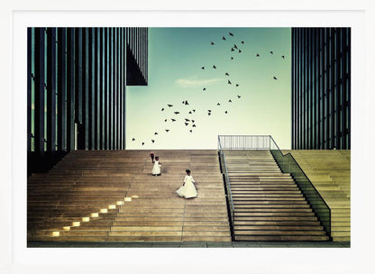 A framed piece of art depicting two young girls in white dresses climbing a vast, modern concrete staircase flanked by two towering, dark buildings. A large flock of birds flies through the teal sky above them, adding a sense of movement to the structured, architectural scene. Poster