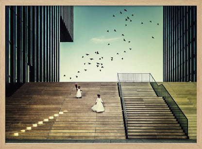 A framed piece of art depicting two young girls in white dresses climbing a vast, modern concrete staircase flanked by two towering, dark buildings. A large flock of birds flies through the teal sky above them, adding a sense of movement to the structured, architectural scene. Poster