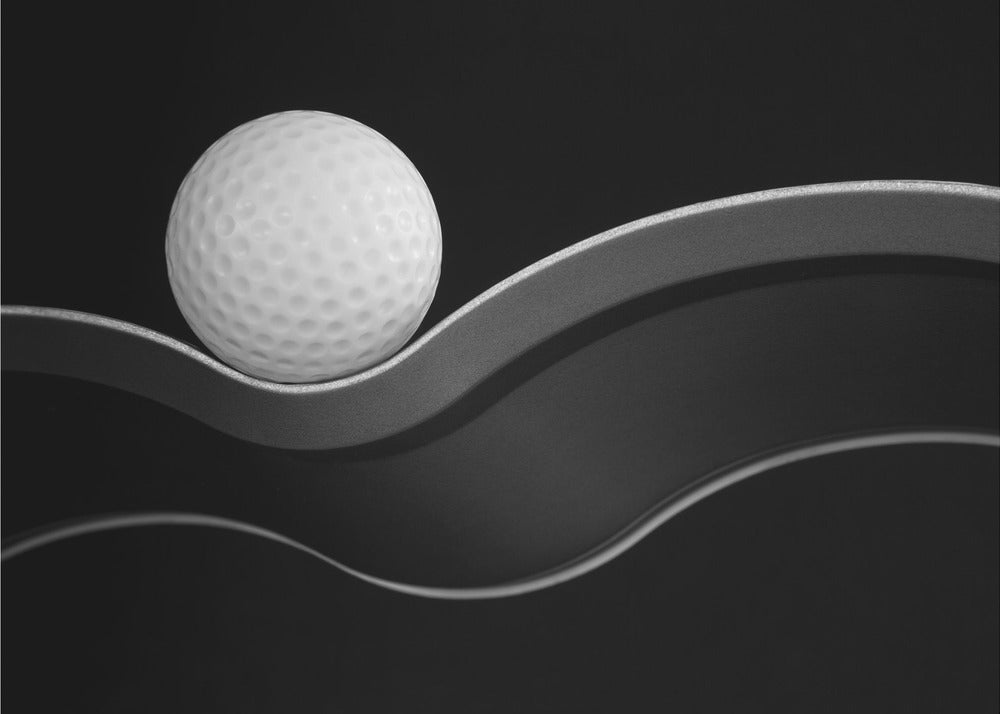 A framed, minimalist black and white photograph of a golf ball resting on a gracefully curving gray surface against a solid black background. Wall Art