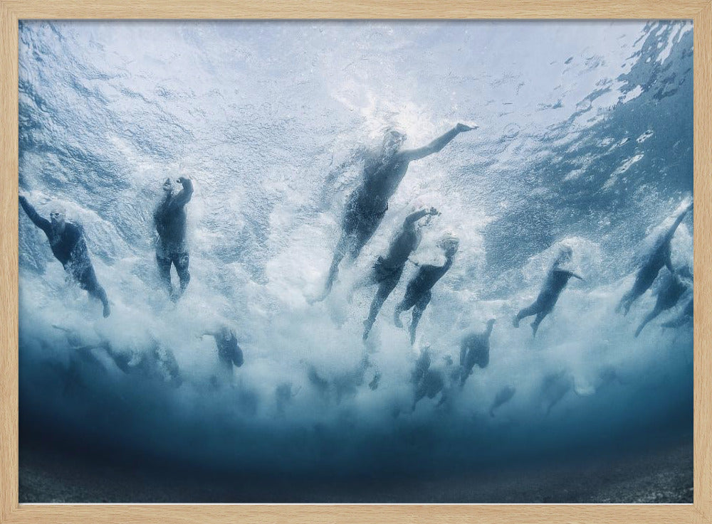 An underwater photograph looking up at a group of swimmers competing in a race. Their bodies are silhouetted against the bright surface of the turbulent blue water, captured mid-motion amidst a flurry of bubbles and foam. Print