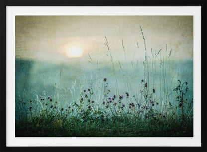 A framed photograph of a misty meadow at sunrise. The soft, pale sun hangs in a hazy, textured sky. In the foreground, tall green grasses and small purple wildflowers are in soft focus, creating a serene and dreamy atmosphere. Decor