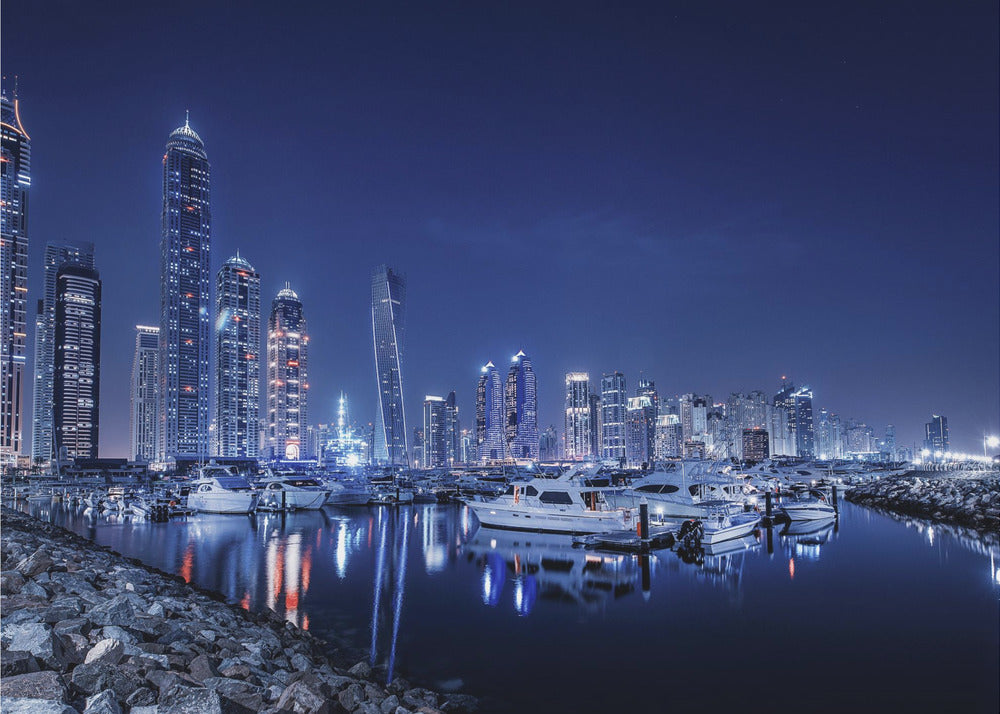 A stunning nighttime photograph of the Dubai Marina skyline, with illuminated skyscrapers towering against a deep blue sky. In the foreground, luxury yachts are moored in the calm harbor, their white hulls and the city's lights creating vibrant reflections on the water's surface. The shot is taken from a rocky embankment. Decor