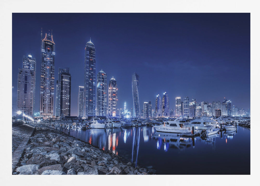A stunning nighttime photograph of the Dubai Marina skyline, with illuminated skyscrapers towering against a deep blue sky. In the foreground, luxury yachts are moored in the calm harbor, their white hulls and the city's lights creating vibrant reflections on the water's surface. The shot is taken from a rocky embankment. Decor