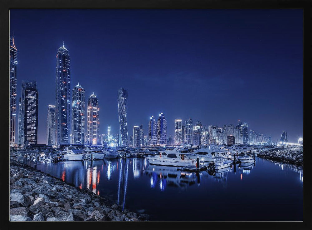 A stunning nighttime photograph of the Dubai Marina skyline, with illuminated skyscrapers towering against a deep blue sky. In the foreground, luxury yachts are moored in the calm harbor, their white hulls and the city's lights creating vibrant reflections on the water's surface. The shot is taken from a rocky embankment. Decor