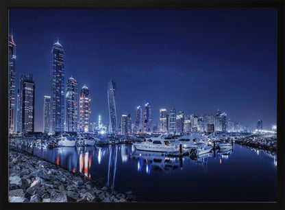 A stunning nighttime photograph of the Dubai Marina skyline, with illuminated skyscrapers towering against a deep blue sky. In the foreground, luxury yachts are moored in the calm harbor, their white hulls and the city's lights creating vibrant reflections on the water's surface. The shot is taken from a rocky embankment. Decor