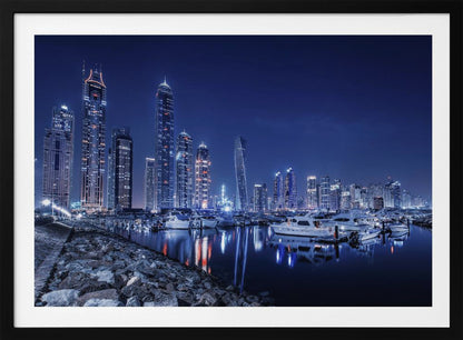 A stunning nighttime photograph of the Dubai Marina skyline, with illuminated skyscrapers towering against a deep blue sky. In the foreground, luxury yachts are moored in the calm harbor, their white hulls and the city's lights creating vibrant reflections on the water's surface. The shot is taken from a rocky embankment. Decor
