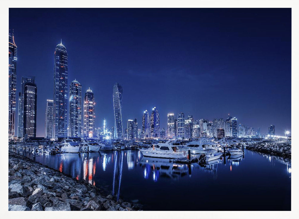 A stunning nighttime photograph of the Dubai Marina skyline, with illuminated skyscrapers towering against a deep blue sky. In the foreground, luxury yachts are moored in the calm harbor, their white hulls and the city's lights creating vibrant reflections on the water's surface. The shot is taken from a rocky embankment. Decor