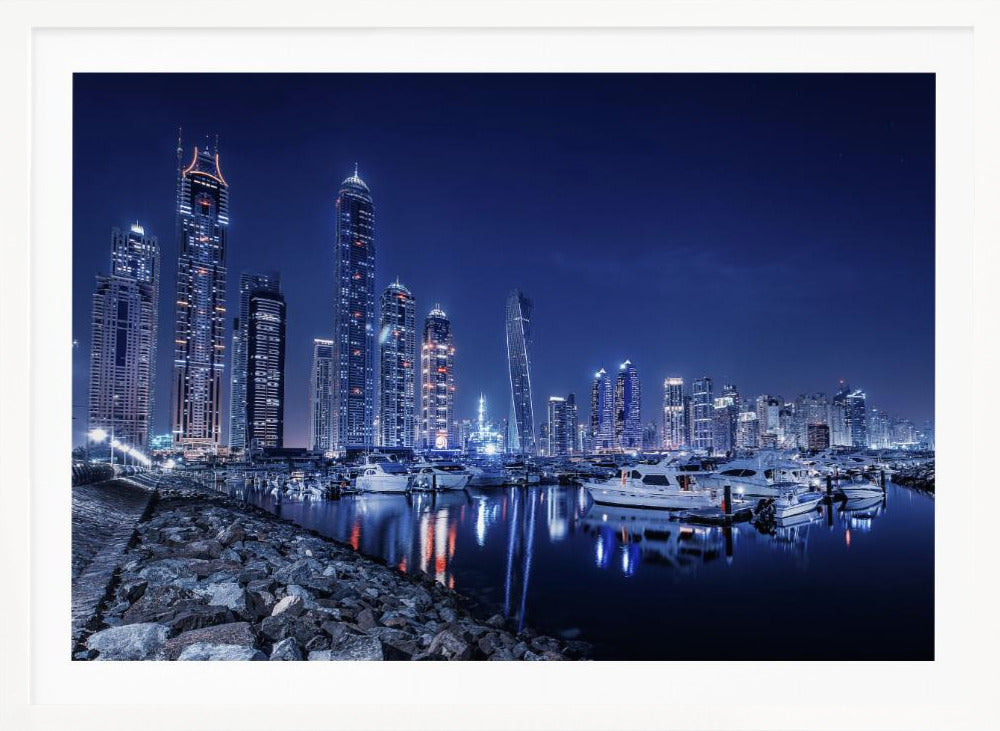 A stunning nighttime photograph of the Dubai Marina skyline, with illuminated skyscrapers towering against a deep blue sky. In the foreground, luxury yachts are moored in the calm harbor, their white hulls and the city's lights creating vibrant reflections on the water's surface. The shot is taken from a rocky embankment. Decor