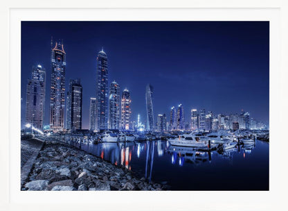 A stunning nighttime photograph of the Dubai Marina skyline, with illuminated skyscrapers towering against a deep blue sky. In the foreground, luxury yachts are moored in the calm harbor, their white hulls and the city's lights creating vibrant reflections on the water's surface. The shot is taken from a rocky embankment. Decor