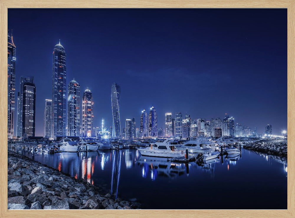 A stunning nighttime photograph of the Dubai Marina skyline, with illuminated skyscrapers towering against a deep blue sky. In the foreground, luxury yachts are moored in the calm harbor, their white hulls and the city's lights creating vibrant reflections on the water's surface. The shot is taken from a rocky embankment. Decor
