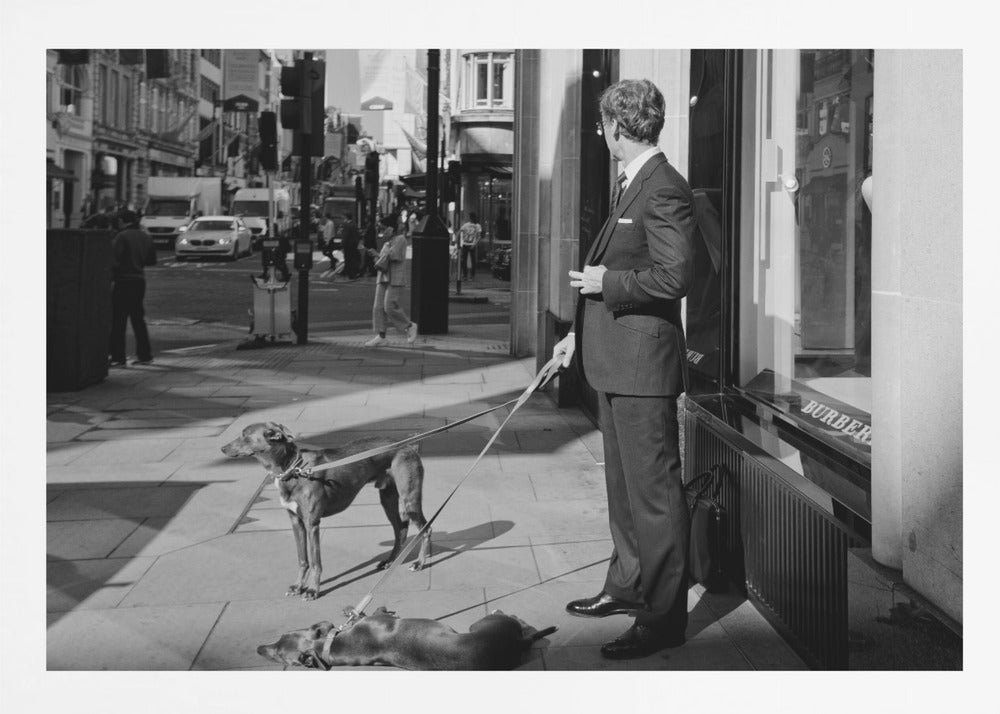 A black and white street photograph of a well-dressed man in a suit standing on a city sidewalk outside a storefront. He holds the leashes for two dogs; one is standing alert, and the other is lying on its back on the pavement. The background is a busy city street with traffic and pedestrians. Print