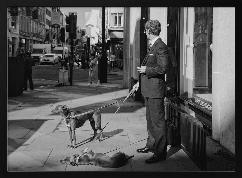 A black and white street photograph of a well-dressed man in a suit standing on a city sidewalk outside a storefront. He holds the leashes for two dogs; one is standing alert, and the other is lying on its back on the pavement. The background is a busy city street with traffic and pedestrians. Print