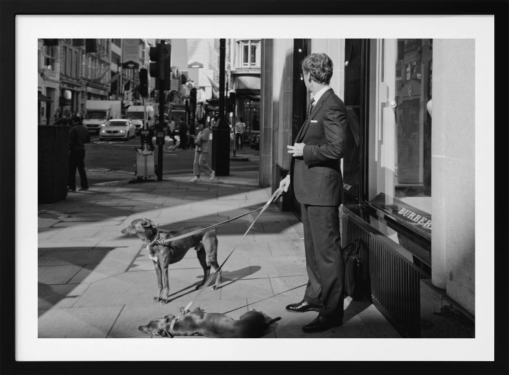 A black and white street photograph of a well-dressed man in a suit standing on a city sidewalk outside a storefront. He holds the leashes for two dogs; one is standing alert, and the other is lying on its back on the pavement. The background is a busy city street with traffic and pedestrians. Print
