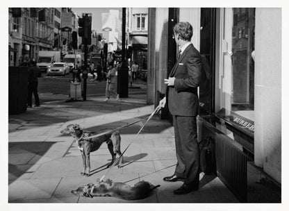A black and white street photograph of a well-dressed man in a suit standing on a city sidewalk outside a storefront. He holds the leashes for two dogs; one is standing alert, and the other is lying on its back on the pavement. The background is a busy city street with traffic and pedestrians. Print