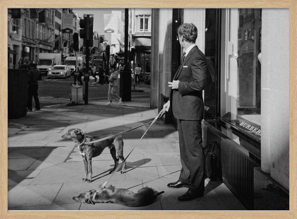 A black and white street photograph of a well-dressed man in a suit standing on a city sidewalk outside a storefront. He holds the leashes for two dogs; one is standing alert, and the other is lying on its back on the pavement. The background is a busy city street with traffic and pedestrians. Print
