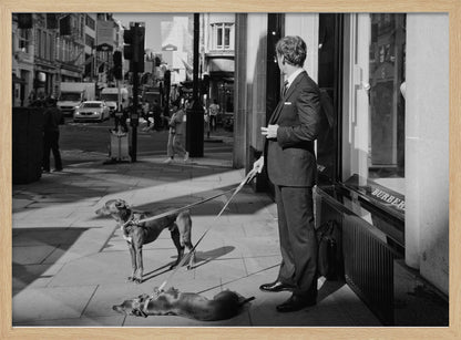 A black and white street photograph of a well-dressed man in a suit standing on a city sidewalk outside a storefront. He holds the leashes for two dogs; one is standing alert, and the other is lying on its back on the pavement. The background is a busy city street with traffic and pedestrians. Print