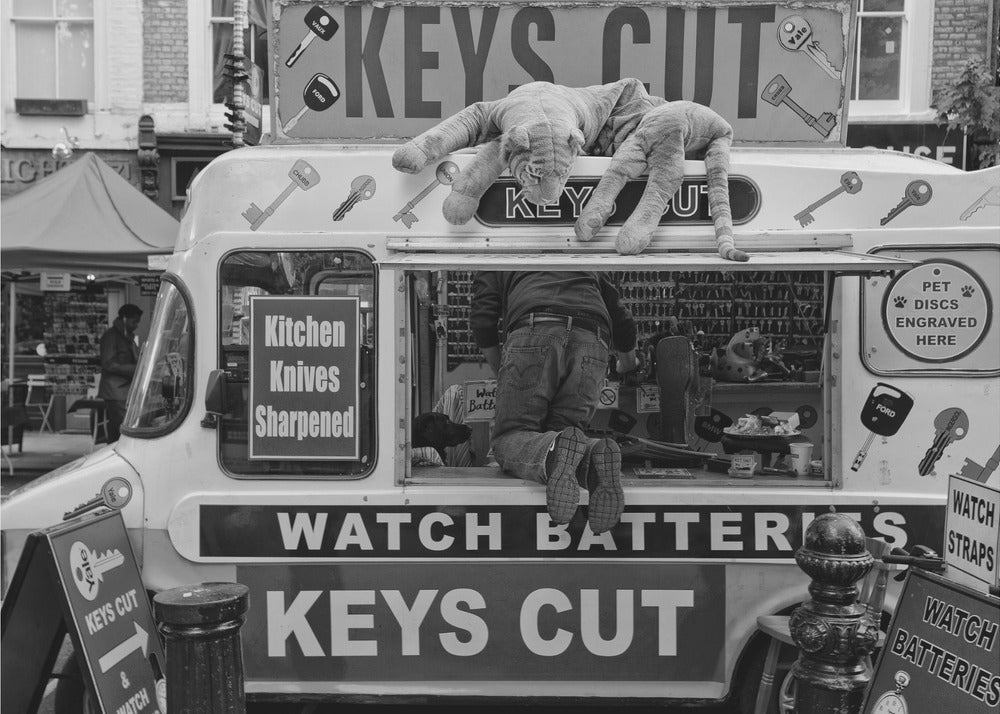 A quirky black and white photograph of a mobile key-cutting van on a city street. A man in jeans is seen from behind, kneeling inside the service window. On the roof, a large stuffed tiger lies stretched out, peering down. The van is covered in signs advertising 'KEYS CUT', 'WATCH BATTERIES', and 'Kitchen Knives Sharpened'. Print